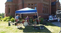 Photo of three men playing musical instruments under a blue canopy in front of a brick building