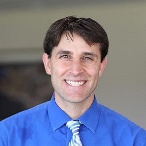 Photo of a smiling man with dark hair and a blue shirt and striped tie