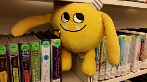 Photo of a yellow stuffed animal monster on a shelf with library audiobooks