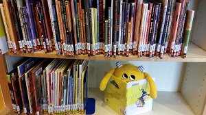 Photo of a yellow stuffed animal monster sitting on a library shelf reading a book