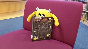 Photo of a yellow stuffed animal monster sitting on a purple chair reading a book