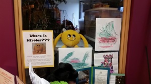 Photo of a yellow stuffed animal monster sitting in the window to an office