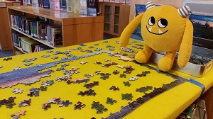 Photo of a yellow stuffed animal monster sitting by a jigsaw puzzle