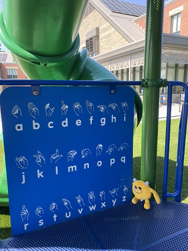 Nibbles the yellow book monster sitting on a blue playground structure next to a sign with the sign language alphabet.