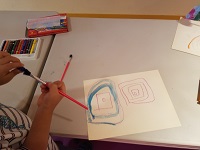 Photo of a child's hands holding two paintbrushes and a table with a piece of paper and art supplies
