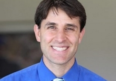 Photo of a smiling man with dark hair and a blue shirt and striped tie
