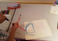 Photo of a child's hands holding two paintbrushes and a table with a piece of paper and art supplies