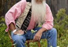 Photo of a man with a long beard wearing a pink shirt multicolored vest and blue jeans